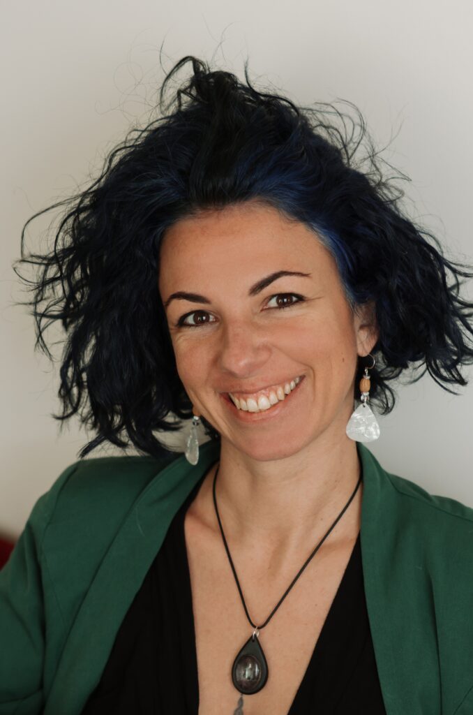 Ambre Louis, psychanalyste, souriant face caméra dans une veste verte avec des cheveux bleus et des boucles d'oreille en noyaux d'olives, médiator et corde de guitar.